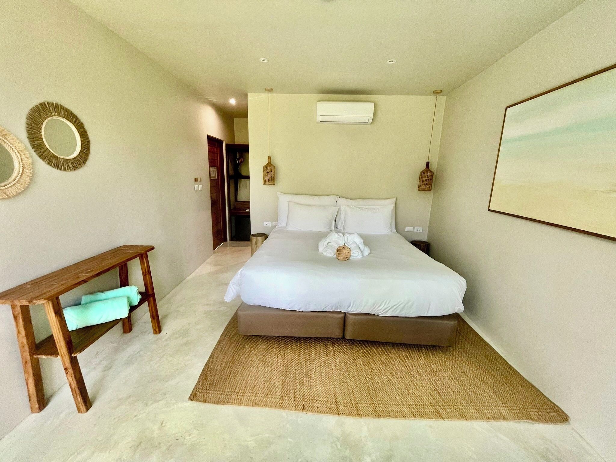 A bedroom with a bed, a bench, a wall-mounted air conditioner, and a door leading to another room.