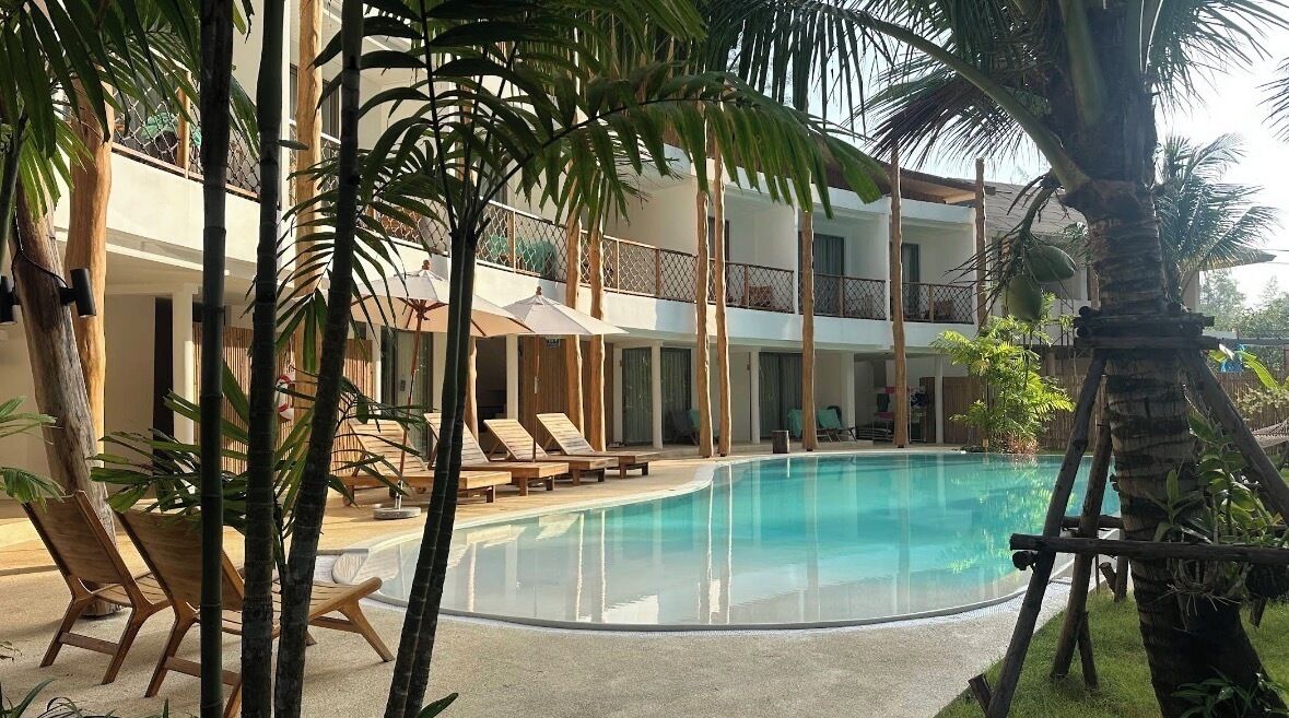A poolside area with lounge chairs, palm trees, and a building with balconies.