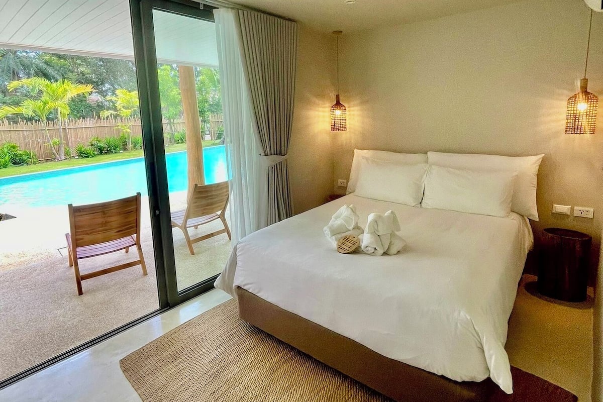 Ein modernes Hotelzimmer mit einem großen Bett, Blick auf einen Swimmingpool und einem Balkon mit einem Stuhl.