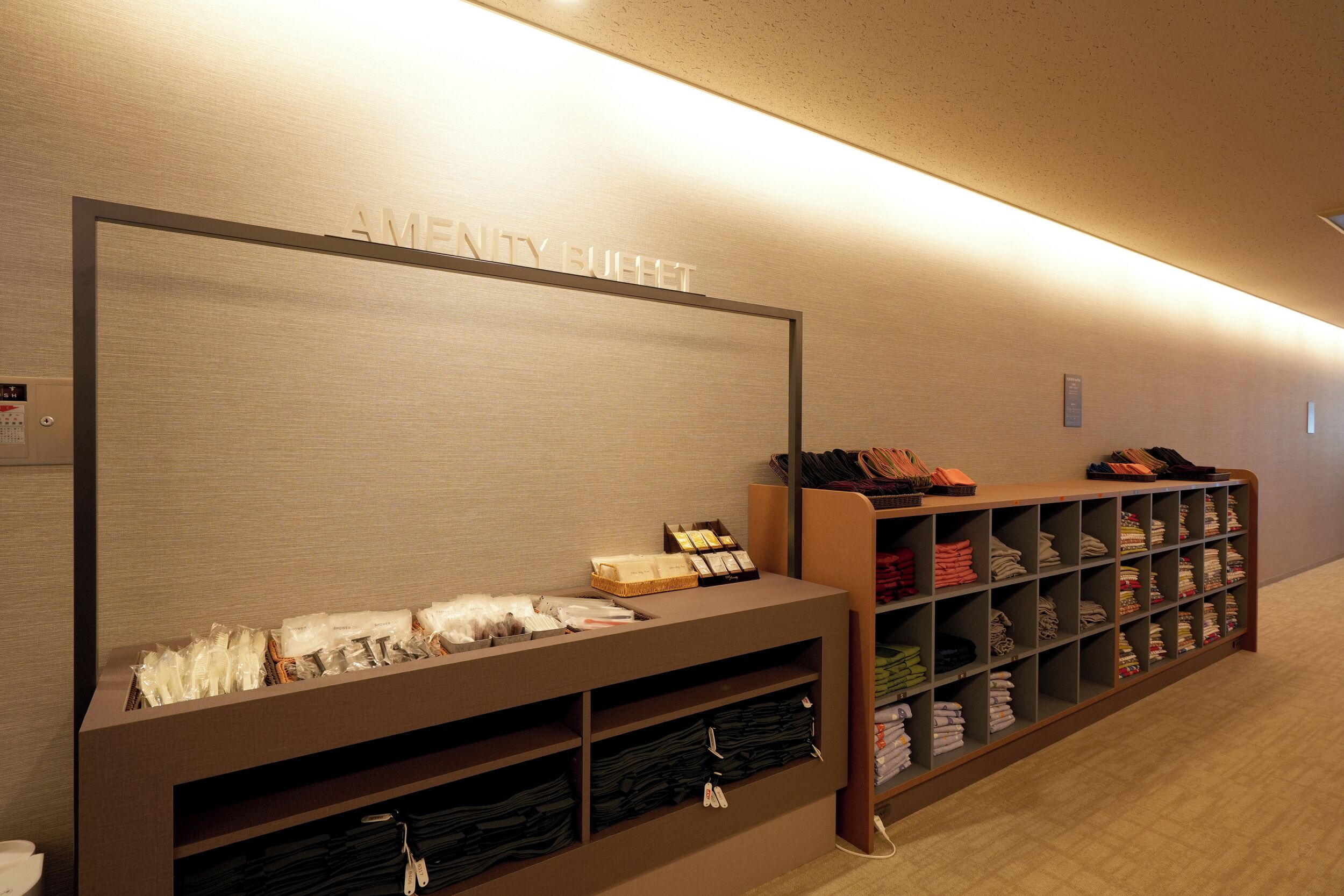 A hotel lobby with a reception desk, a wall-mounted sign reading "AMENITY BOUFFET," and neatly organized shelves with folded towels and other amenities.