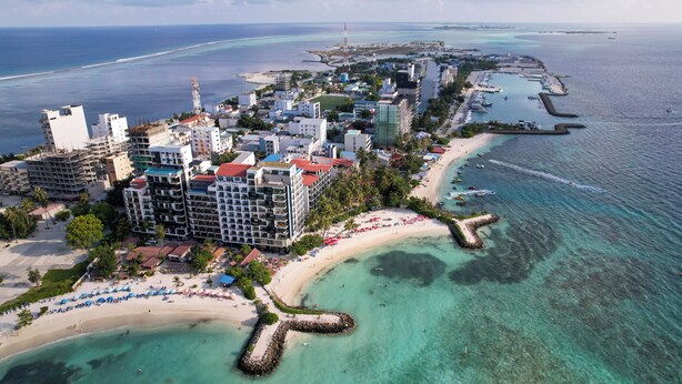 An aerial view of a coastal city with a beachfront hotel, clear turquoise waters, and a rocky breakwater. - SEASUNBEACH MAAFUSHI (Maafushi)