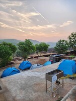 Un camping con varias tiendas azules, una parrilla metálica y vistas a colinas distantes.