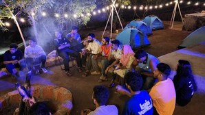 A group of people gathered around a fire pit at night, with tents and string lights in the background. - Aaroha Hill Top (Gokarna)