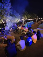 Un grupo de personas reunidas alrededor de una fogata por la noche, con tiendas de campaña y luces colgantes al fondo.