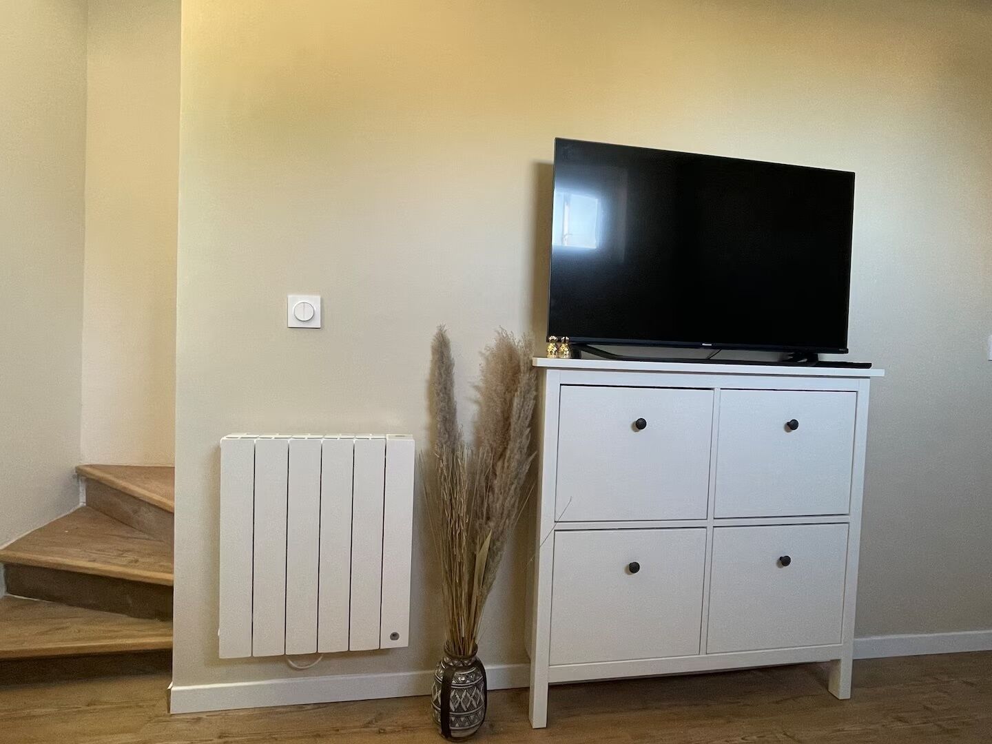 Een kamer met een witte kast, een flatscreen-tv, een radiator en een vaas met gedroogde planten.