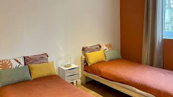 A twin bedroom with two single beds, a nightstand with a lamp, and a window with curtains.