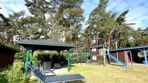 A covered outdoor seating area with chairs and a table, a small wooden playhouse with a slide, and a fenced play area with a swing set. - Relax in the vacation apartment "Sünnhuus" at the coastal forest in Koserow - Am (Koserow)