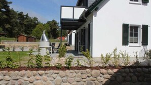 A white building with a stone retaining wall, a chimney, and a small garden area with plants and a metal structure. - Relax in the vacation apartment "Sünnhuus" at the coastal forest in Koserow - Am (Koserow)