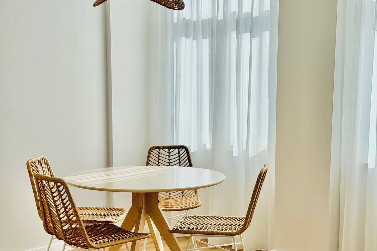 Un comedor con una mesa redonda y cuatro sillas de mimbre, una lámpara colgante y cortinas translúcidas que dejan entrar luz natural.