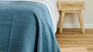 Una cama con cabecera de madera, almohadas blancas y una manta azul con textura. Se observa una mesita de noche con estructura de madera y una lámpara pequeña. El cuarto tiene piso de madera y paredes de color claro.