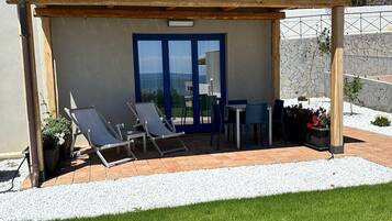 A covered outdoor seating area with a table and chairs, a view of the sea, and a mountain in the background.