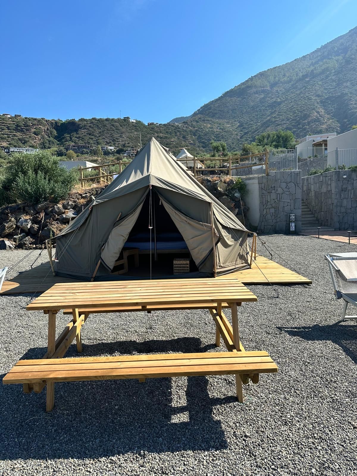 A tent with a picnic table in front, set on a gravel surface with a mountainous backdrop.