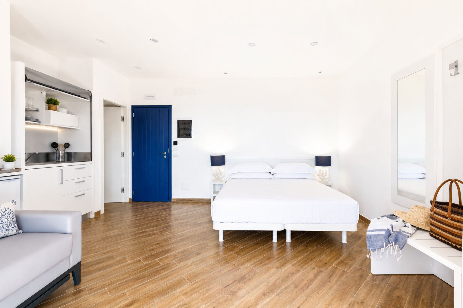 Una habitación de hotel moderna con piso de madera, una cama blanca, una puerta azul, un sofá blanco y un escritorio con una lámpara.