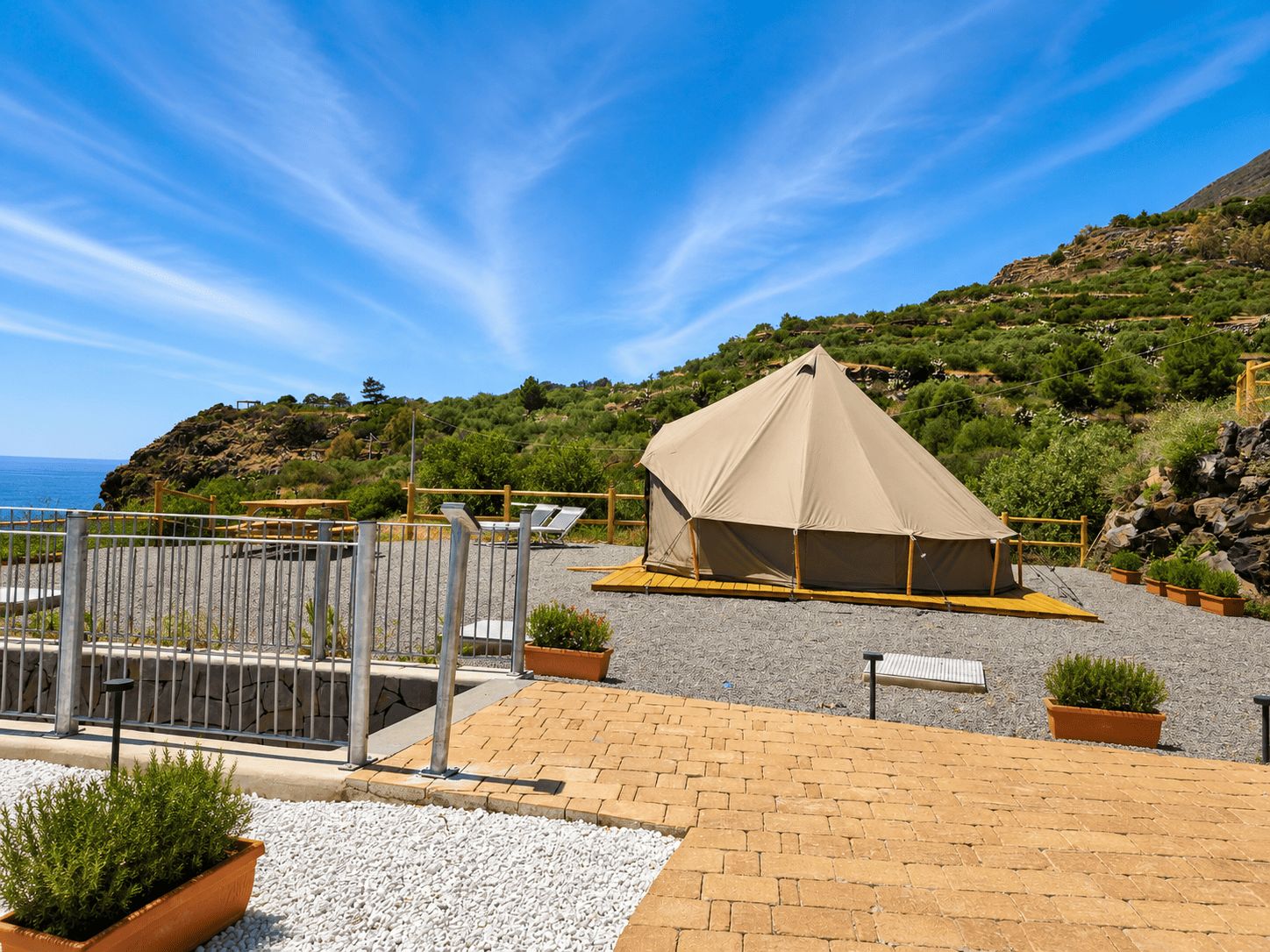 A tented accommodation with a gravel patio, a metal fence, and potted plants.
