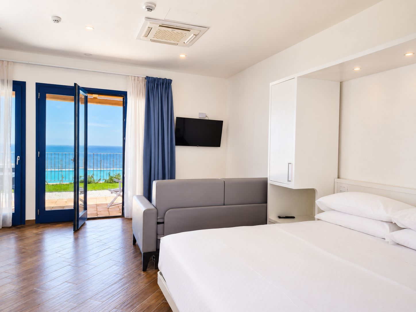 A hotel room with a bed, a sofa, a TV, and a balcony with a view of the ocean.