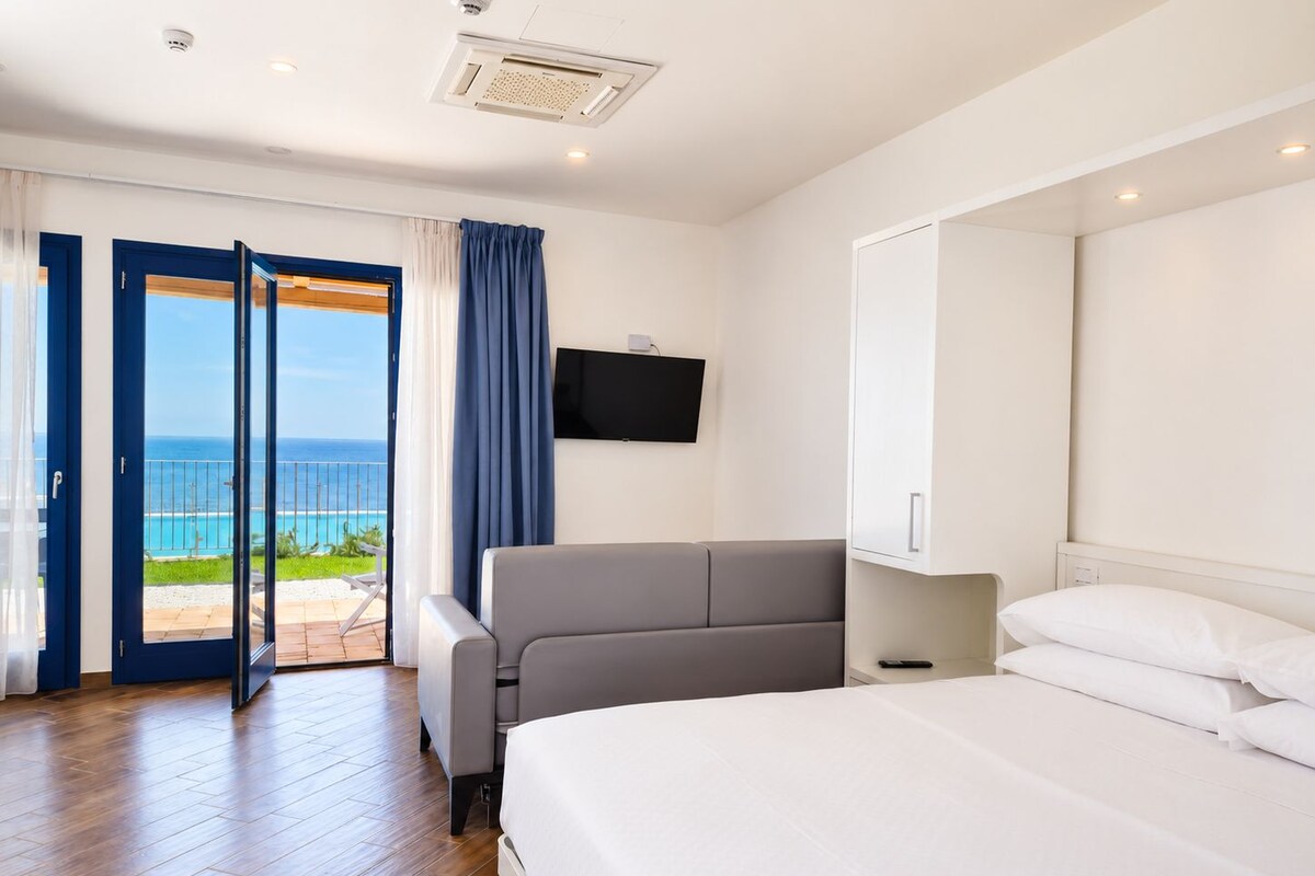 Habitación de hotel con cama, sofá, televisión y balcón con vista al océano.
