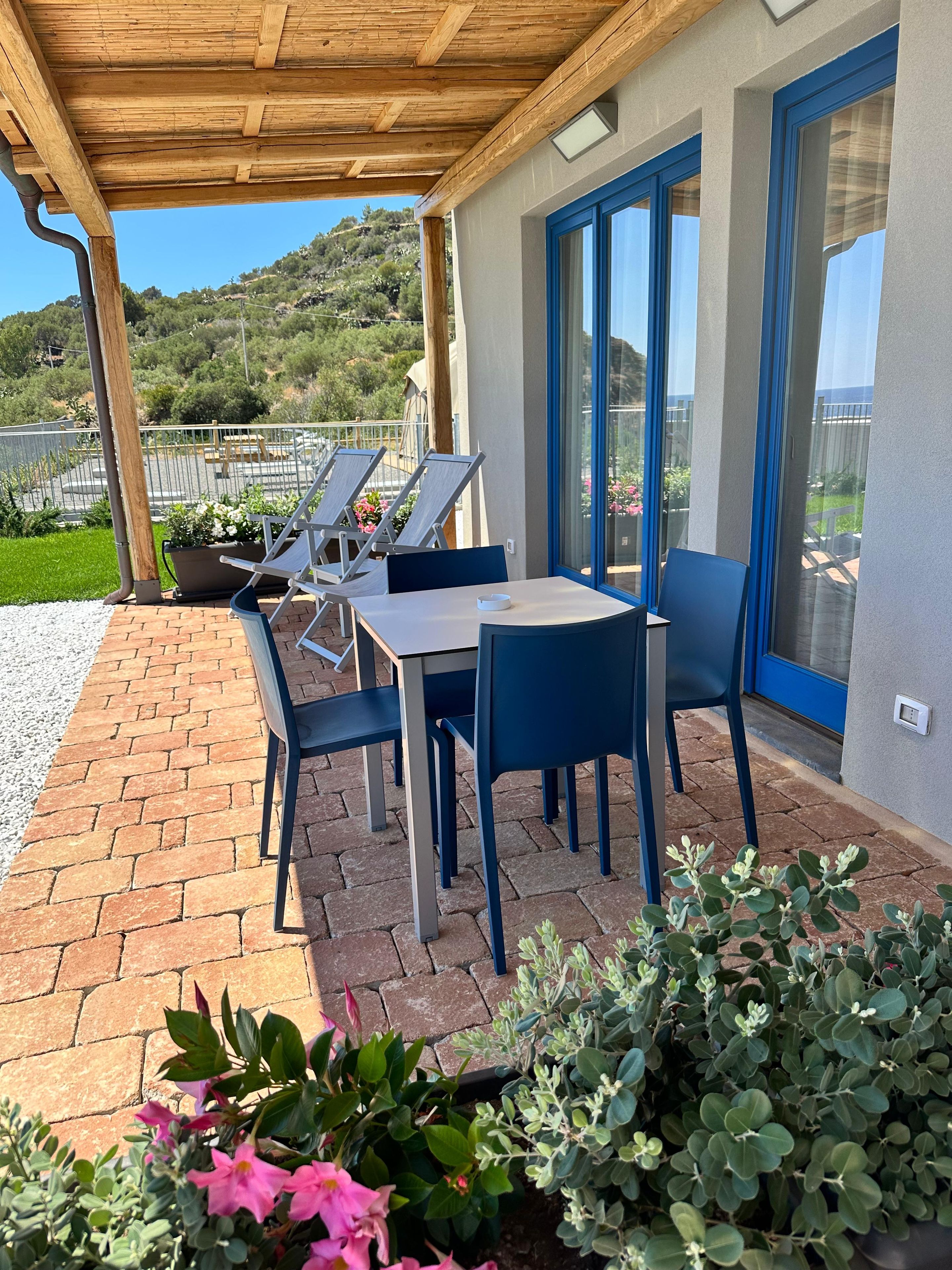 A covered patio with a table and chairs, a view of greenery and hills, and a building with blue-framed windows.