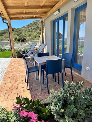Un patio cubierto con una mesa y sillas, vistas a la vegetación y colinas, y un edificio con ventanas de marco azul.