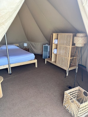 Un alojamiento en carpa con cama, un calentador pequeño, un estante con toallas y una cesta tejida.