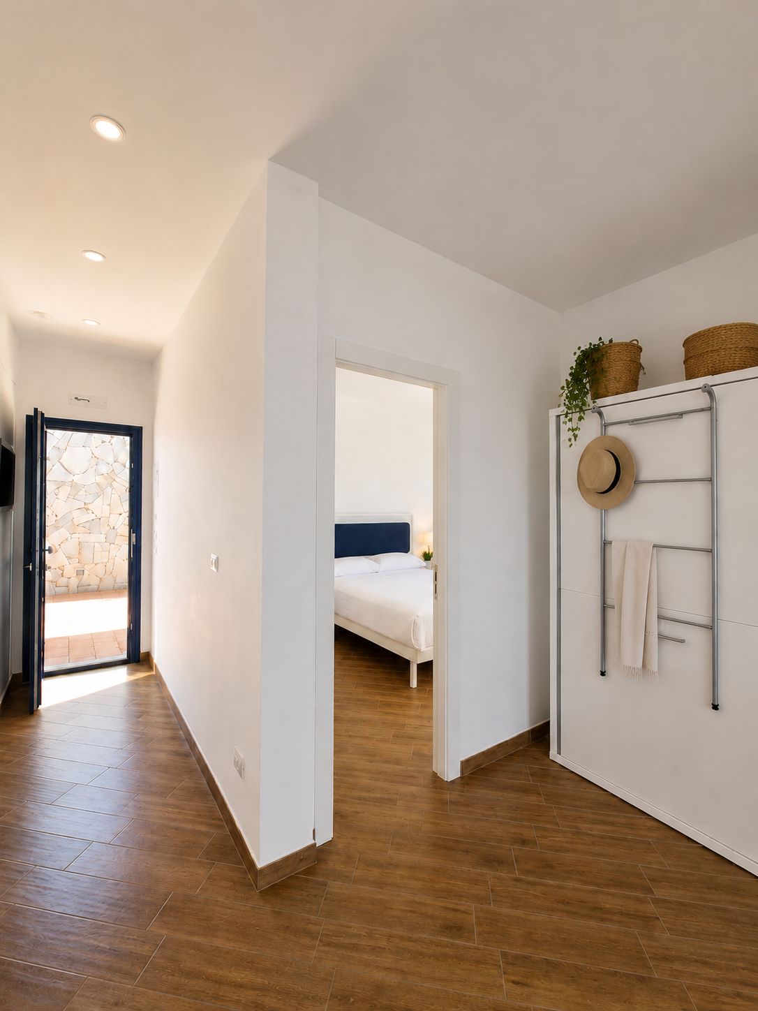 Een hotelkamer met een houten vloer, een bed met witte beddengoed, een wandrek voor handdoeken en een deur naar een balkon.