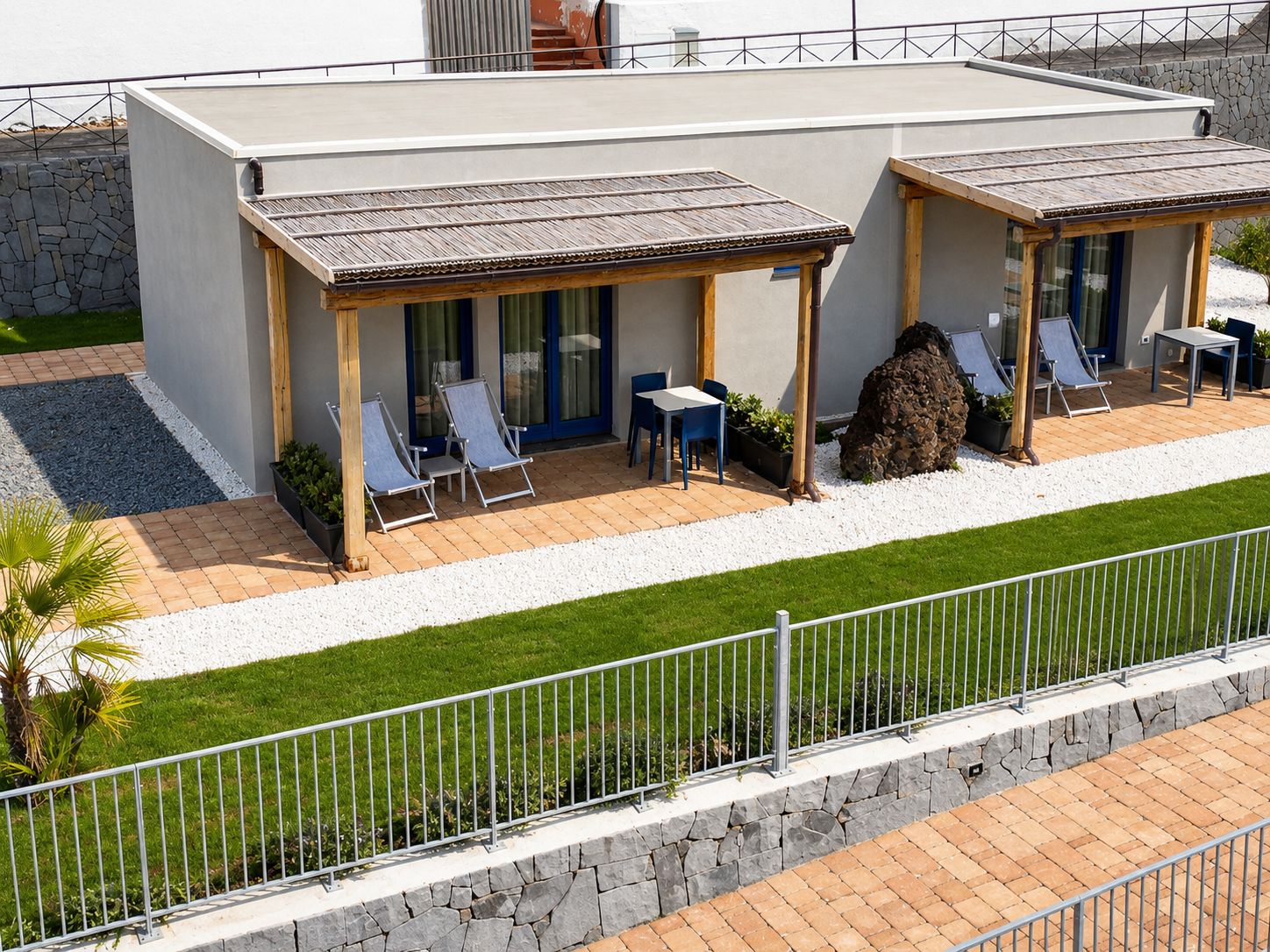 Een modern gebouw met een plat dak, twee overdekte buitenruimtes met stoelen en tafels, en een stenen muur.