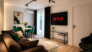 Een moderne woonkamer met een bank, groene stoelen, een salontafel, een televisie waarop 'NETFLIX' staat en een bijzettafeltje met boeken.