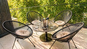 Een ronde tafel met vier zwarte rieten stoelen, een metalen emmer met een fles en verschillende wijnglazen op een balkon met een groene, bladrijke achtergrond.