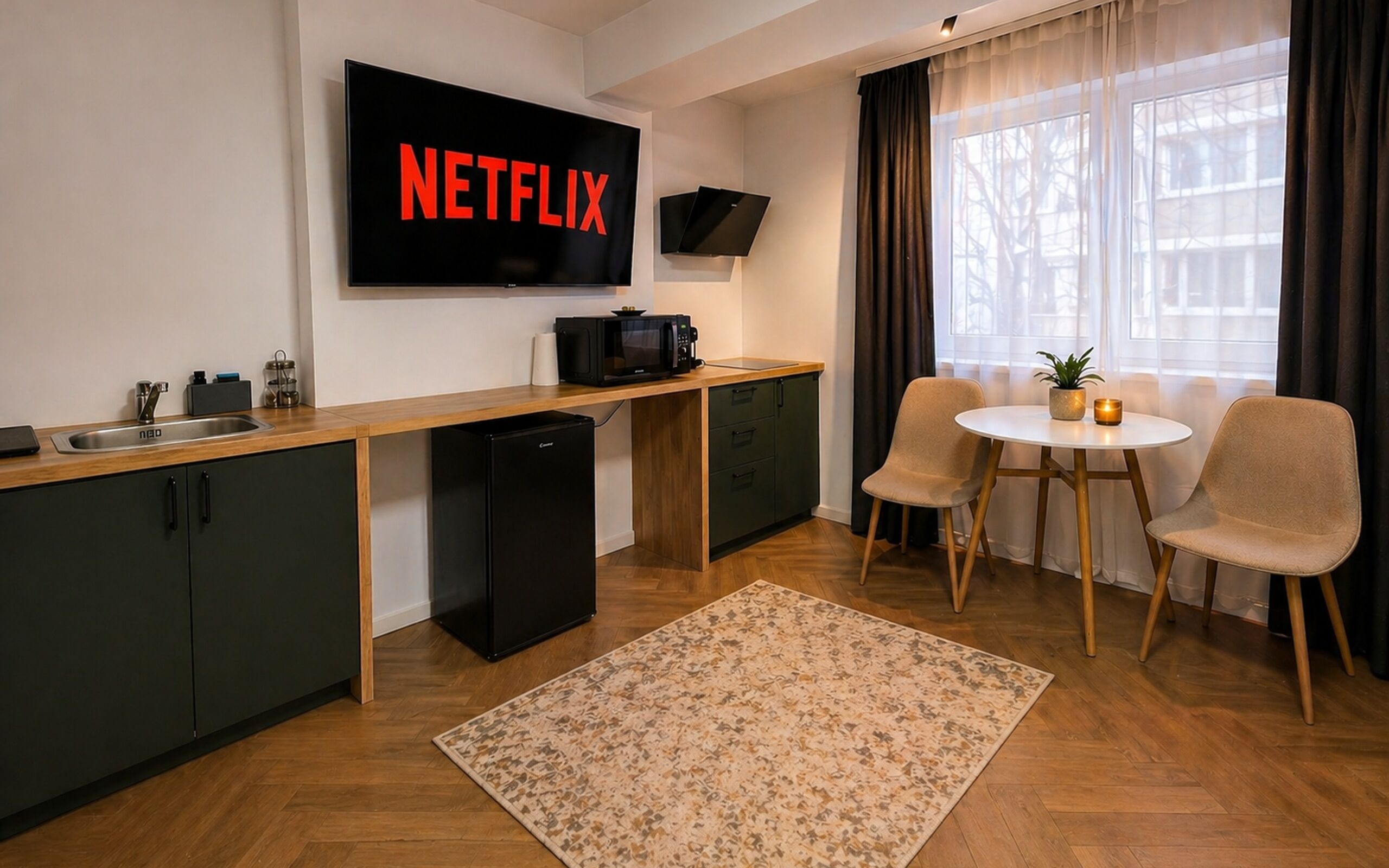 A modern kitchen with a built-in microwave, a sink, and a refrigerator. There is a dining area with two chairs and a small table. A TV mounted on the wall displays the Netflix logo.