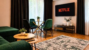 Een moderne woonkamer met een flatscreen waarop 'NETFLIX' staat, een eettafel met glazen blad en stoelen, een groene bank, een klein rond tafeltje met een kaars en een vloerkleed met patroon.