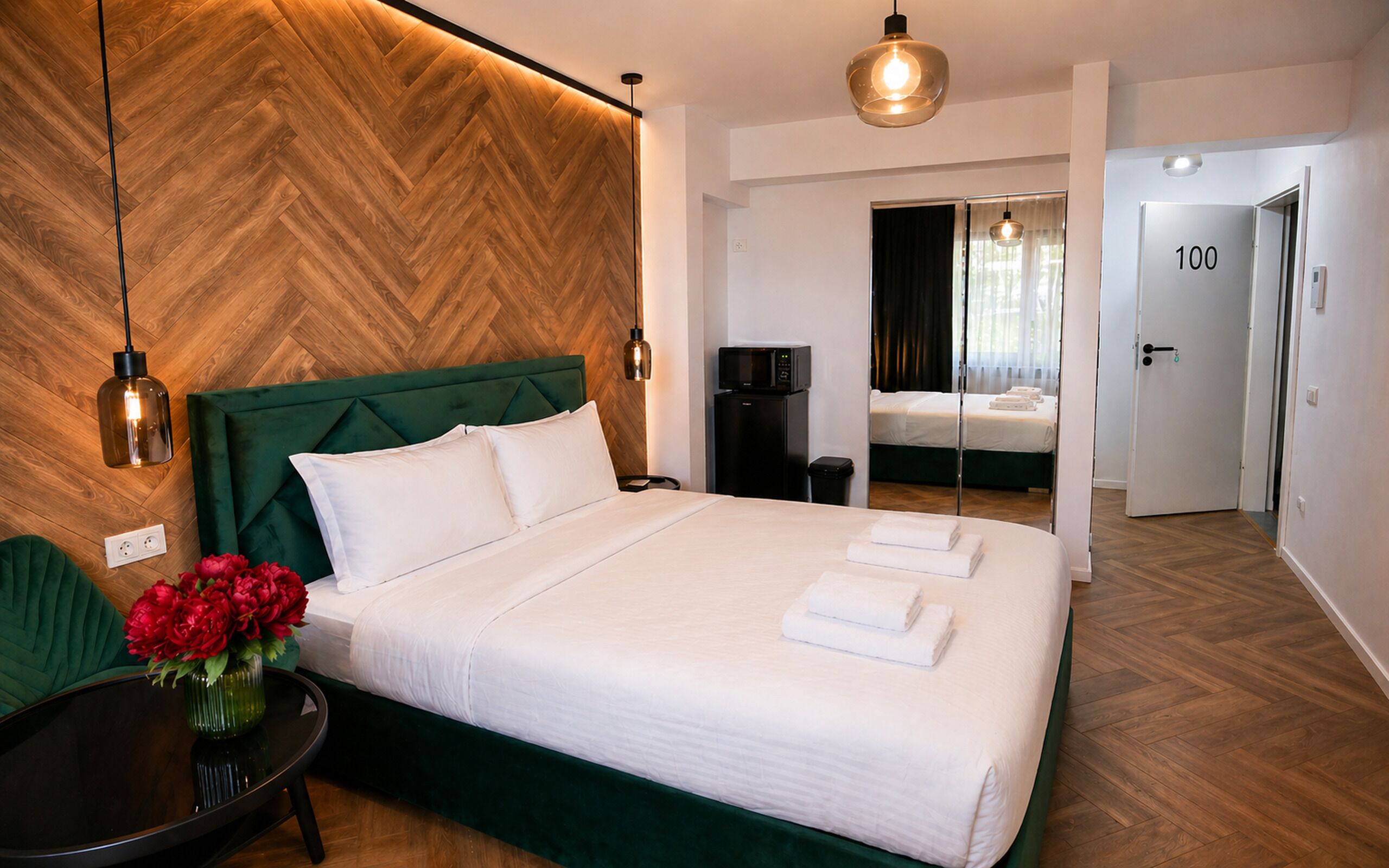 Camera d'albergo con un letto grande, una testiera imbottita verde, un pavimento in legno a spina di pesce, un tavolino con un vaso di fiori e una porta con la scritta '100'.