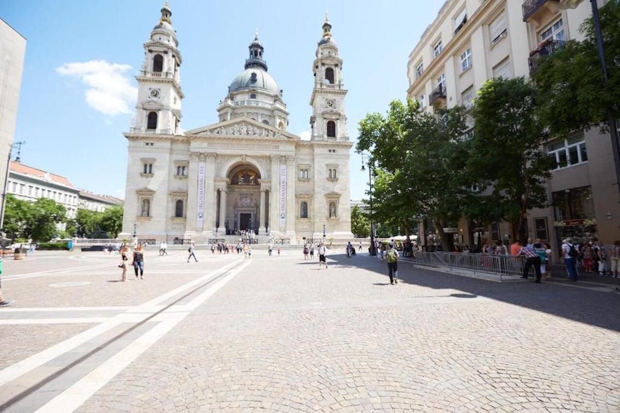 A historic building with a central dome and two towers, surrounded by a paved square with people and trees.