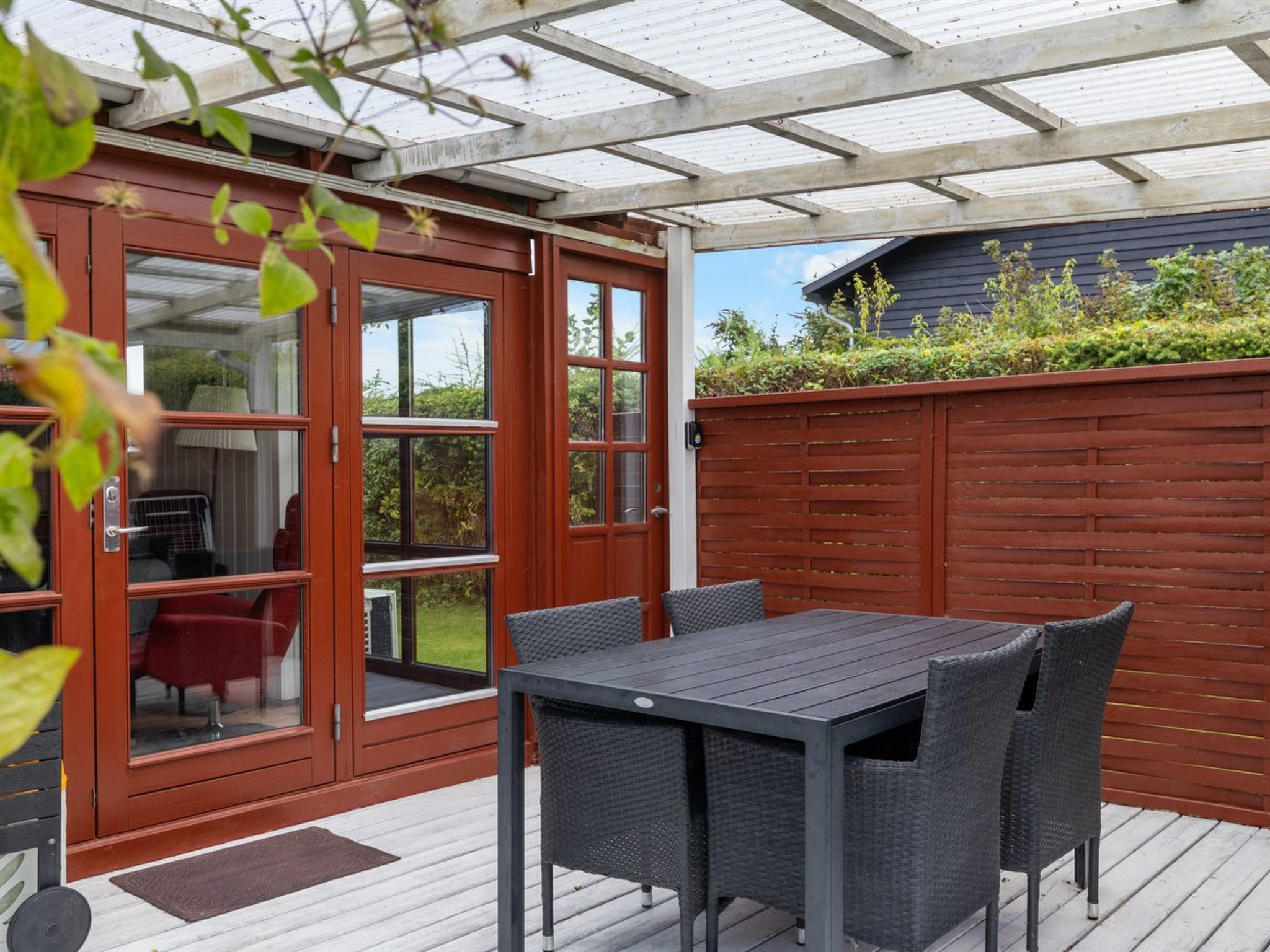 Une terrasse extérieure avec une table et des chaises, une terrasse en bois et une porte vitrée donnant sur une pièce où l’on aperçoit une chaise rouge.