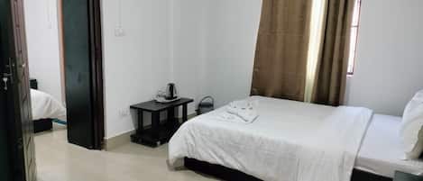 Una habitación de hotel con cama, una mesita con hervidor, maleta y ventana con cortinas.
