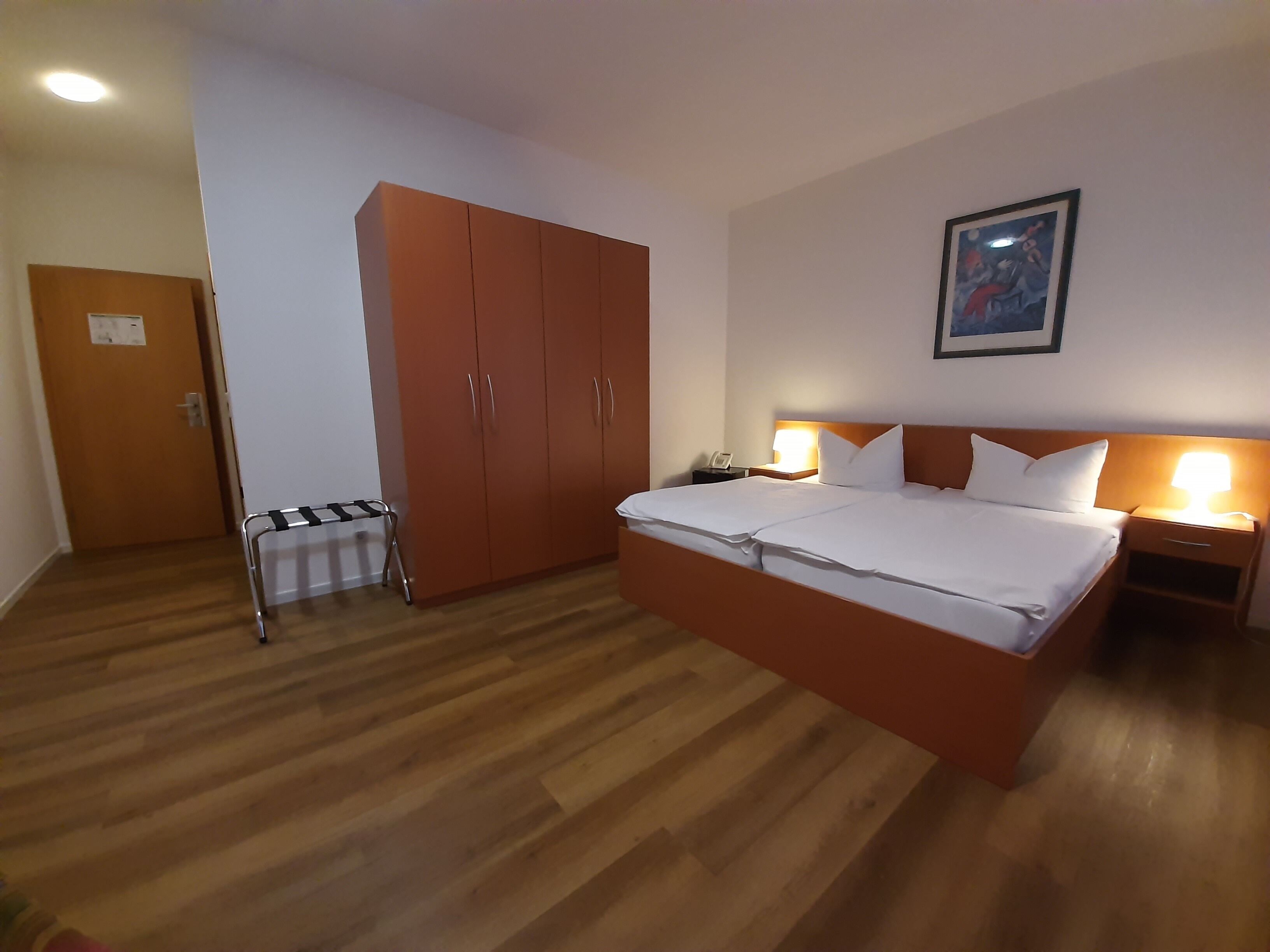 Ein Hotelzimmer mit einem großen Bett, Holzboden, einem Schrank, einem Nachttisch mit Lampe sowie einem Gemälde an der Wand.
