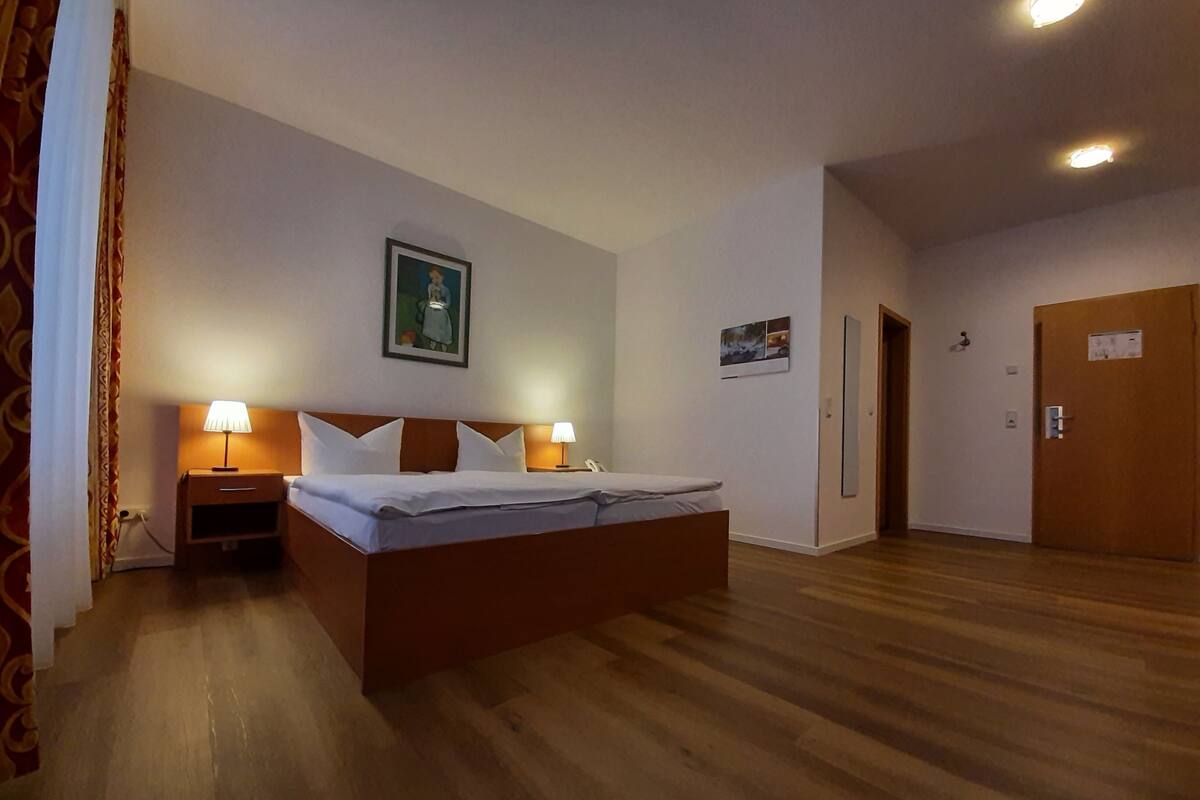 A hotel room with a large bed, two bedside tables with lamps, a wooden headboard, a painting on the wall, and a door to the right.