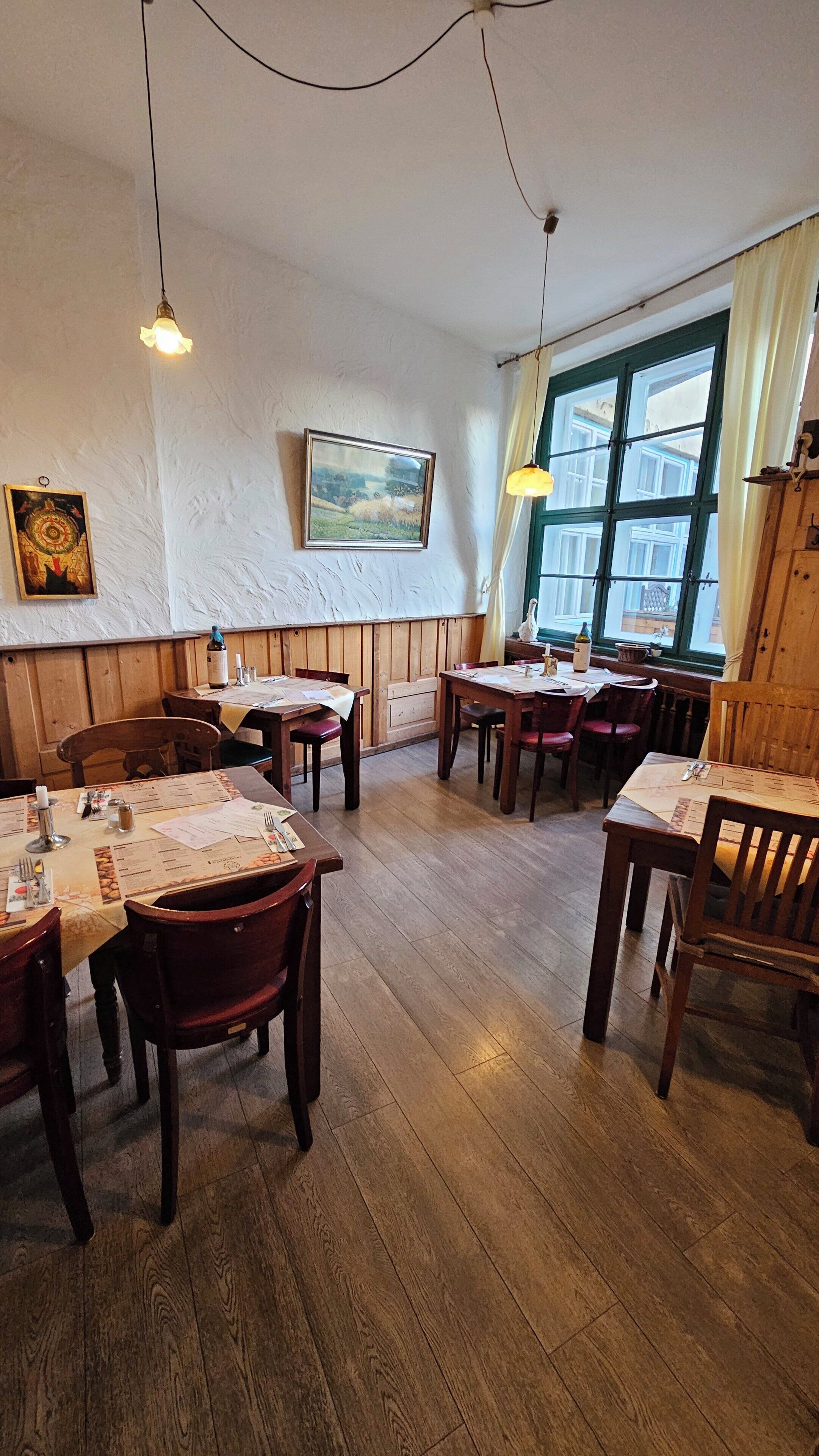 A restaurant with wooden tables and chairs, a large window with curtains, and hanging light fixtures.