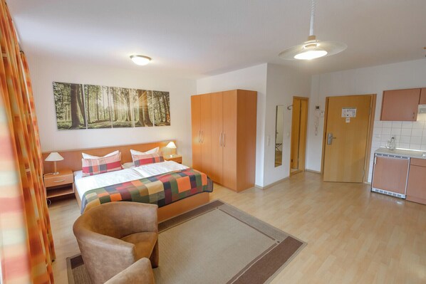 A bedroom with a bed, nightstands, a chair, a large forest-themed painting, and a kitchenette with cabinets.