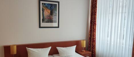A hotel room with a large bed, two bedside tables, a nightstand, a framed picture on the wall, and a window with curtains.