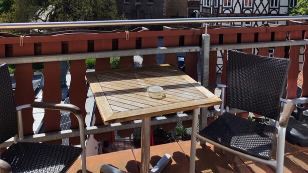 A terrace with a table and chairs, overlooking a historic building with a pointed tower and a traditional house with a gabled roof. - Altwernigeröder Apparthotel  (Wernigerode)