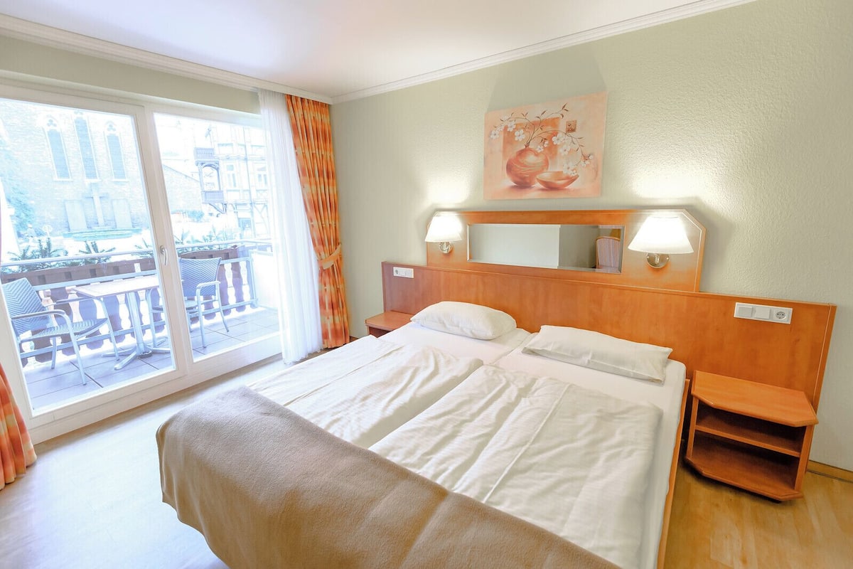 A hotel room with a large bed, a balcony with outdoor seating, and a painting on the wall.