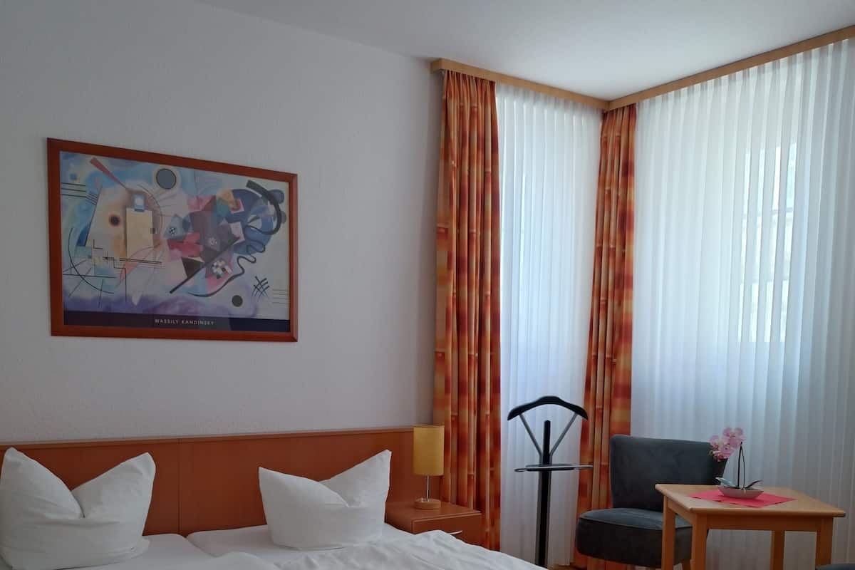 A hotel room with a bed, a chair, a small table, a lamp, and a painting on the wall.