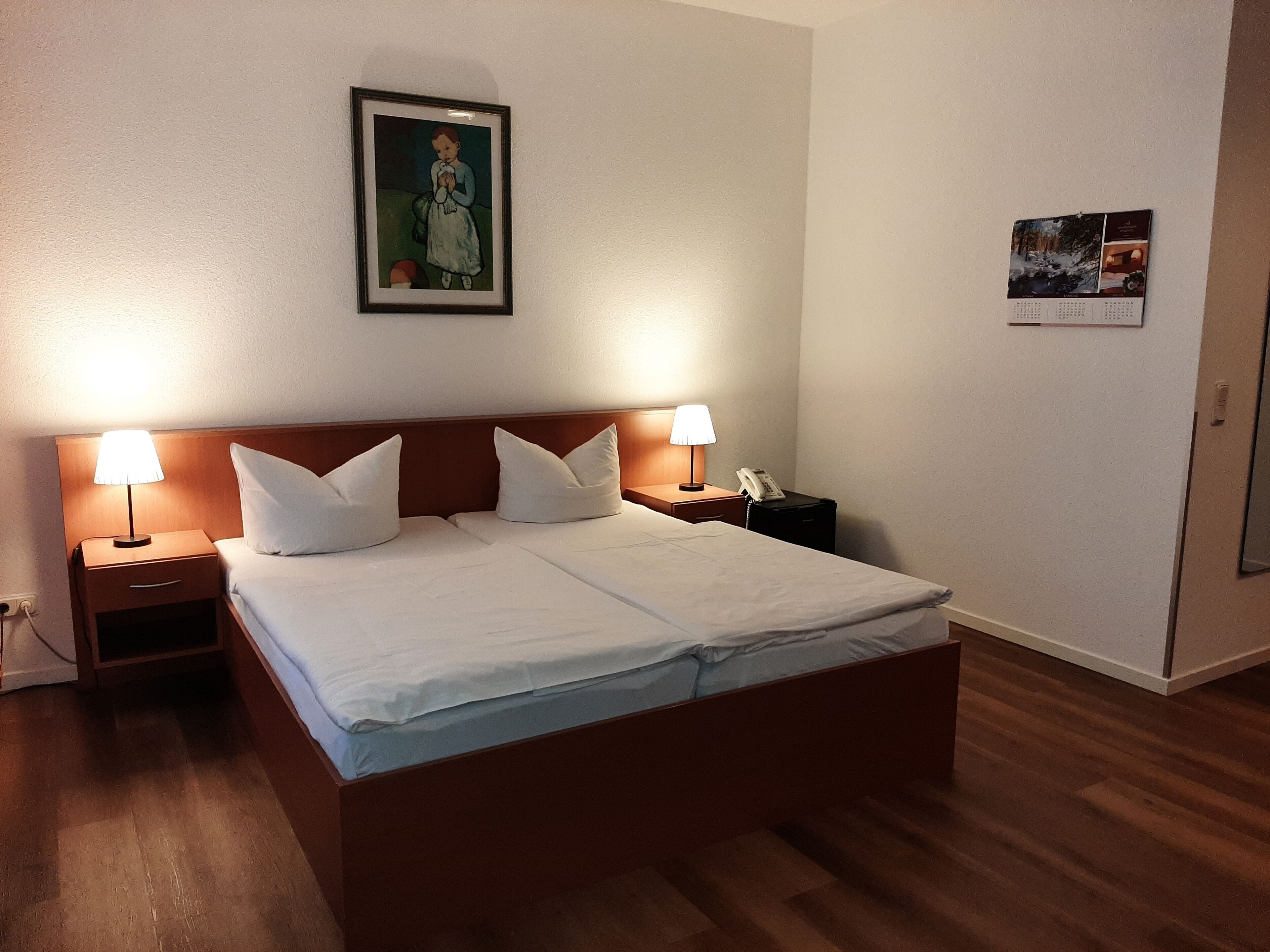 A neatly made bed with white linens, two bedside tables with lamps, a framed picture on the wall, and a calendar.