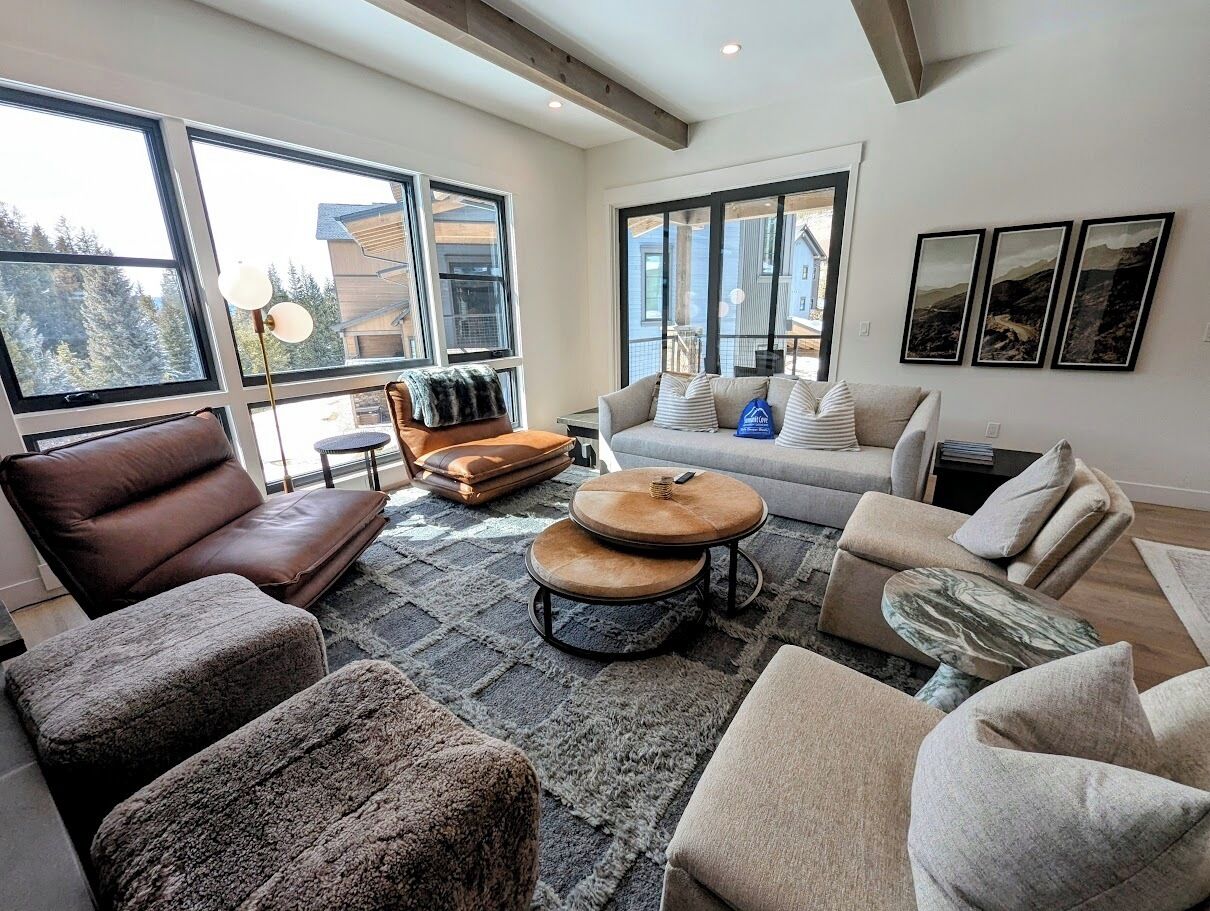 A spacious living room with large windows, a brown leather sofa, a grey sofa, and a round coffee table.
