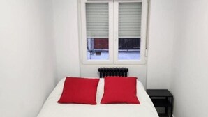Una habitación pequeña y blanca con una cama, dos almohadas rojas, un radiador y una ventana con persianas cerradas.