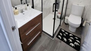 Un baño moderno con ducha envuelta en vidrio, tocador de madera, lavamanos blanco y un inodoro.