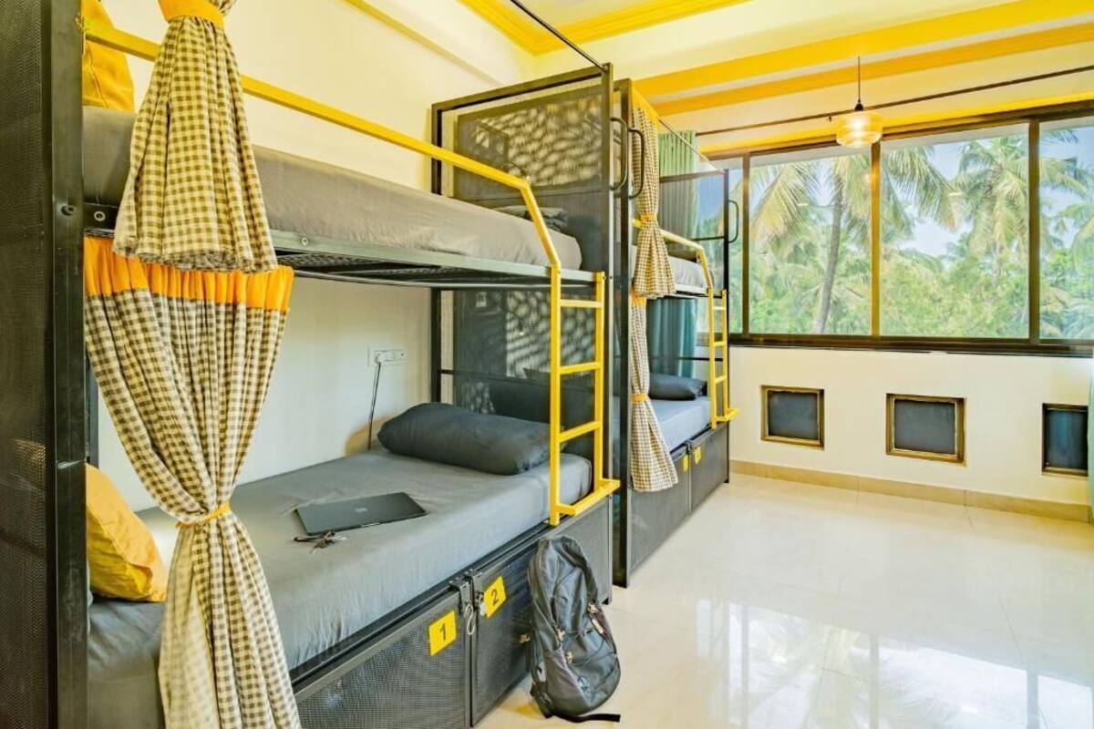 A dormitory room with bunk beds, a window with a view of palm trees, and a bright yellow ceiling.