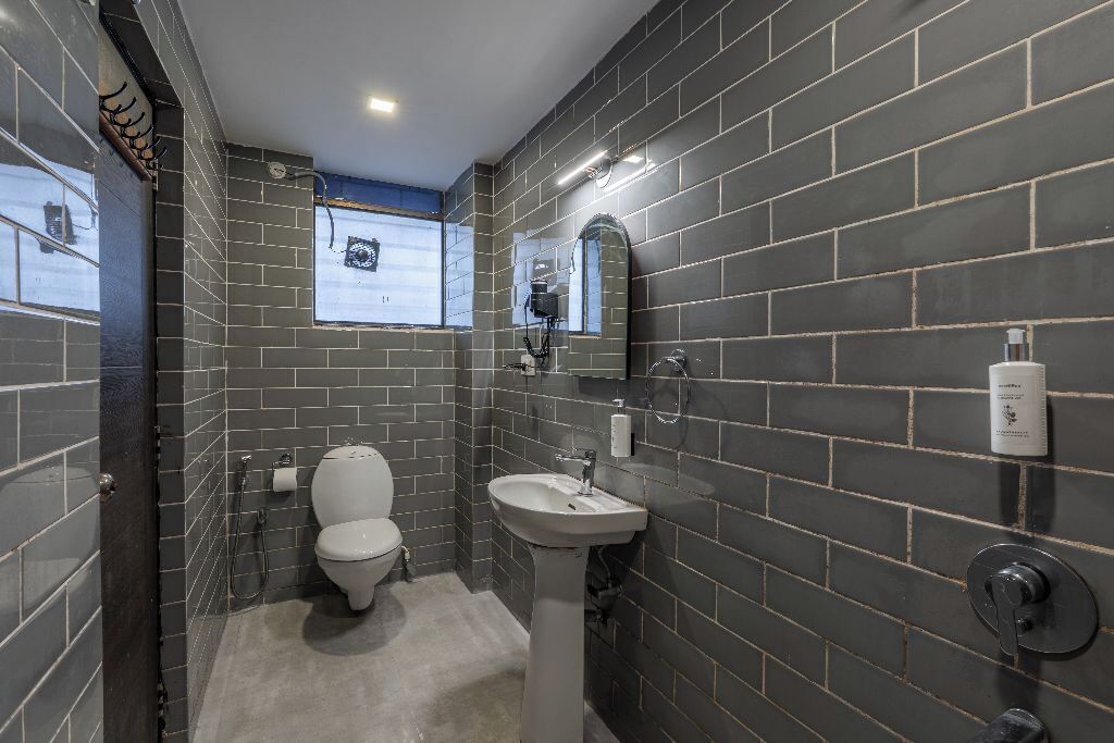 Une salle de bain avec des murs recouverts de carreaux foncés, des toilettes blanches, un lavabo sur colonne et un miroir.