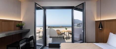 A modern hotel room with a large bed, a desk, and a balcony with a view of the sea.