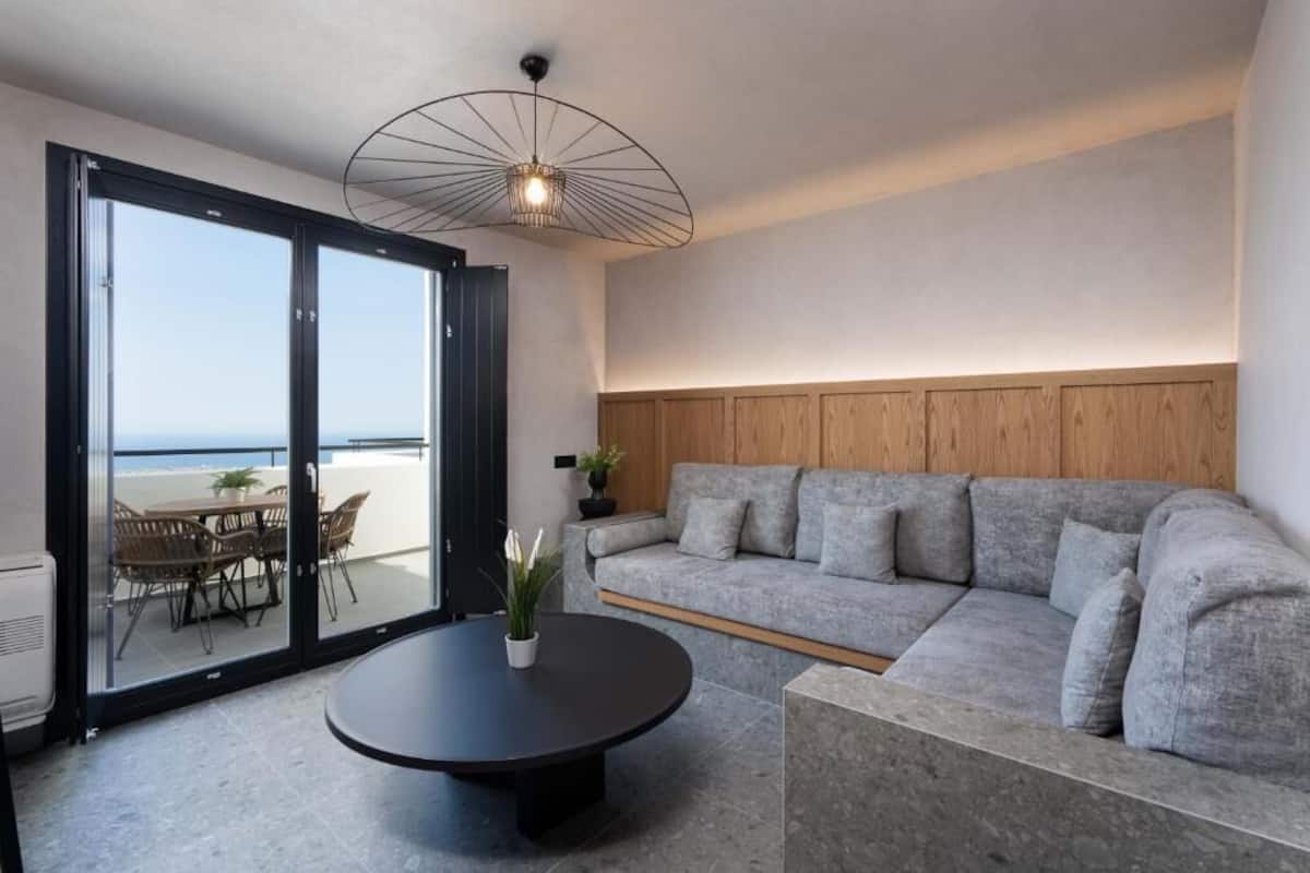 Ein modernes Wohnzimmer mit grauem Sofa, rundem Couchtisch und einem Essbereich mit Stühlen und Tisch. Ein Schiebetür öffnet sich zu einem Balkon mit Meerblick.