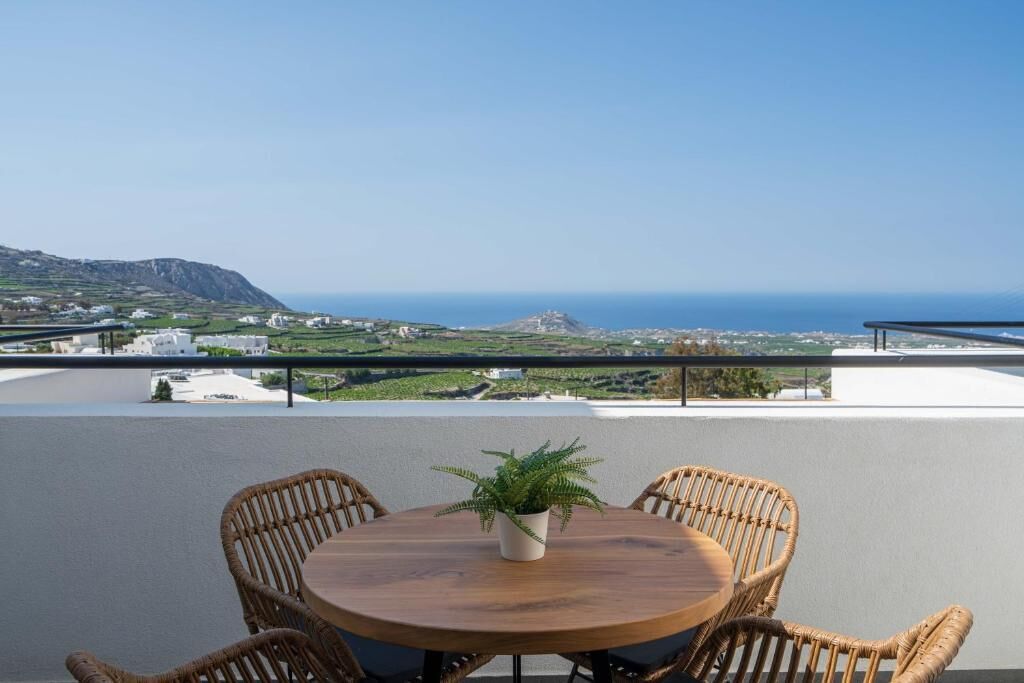 A round wooden table with wicker chairs and a potted plant, overlooking a coastal landscape with buildings and a clear sky.